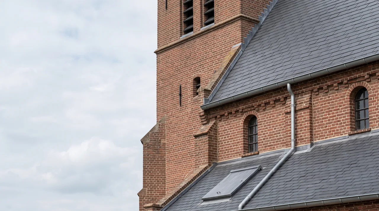 Dakrand van een bakstenen kerk met zinken goot en hemelwaterafvoer, loodslabben tegen het leien dak en een dakluik in het dakvlak.